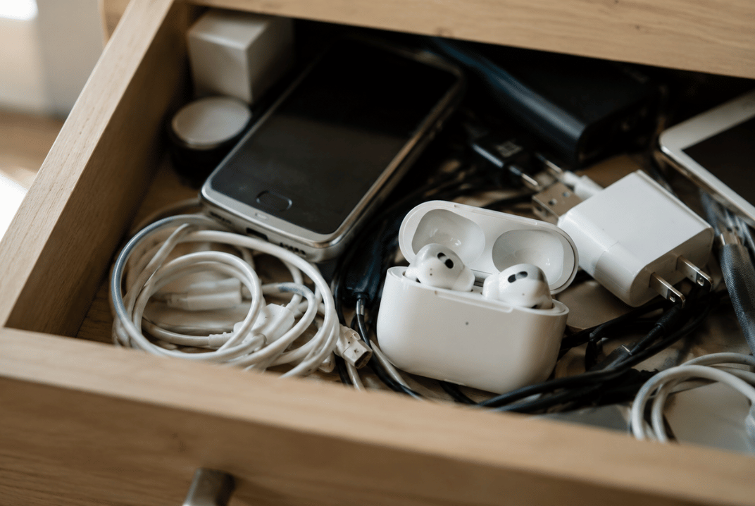 Tangled cables, white wireless earbuds in their charging case, and power adapters inside a wooden drawer, representing common electronic waste that needs proper recycling.